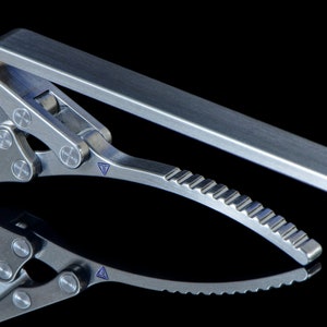 May include: A silver-toned metal tie clip with a unique, multi-jointed design. The clip has a textured, serrated edge and a small, triangular detail on the side.