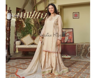 May include: A young person wearing a cream-colored shalwar kameez with gold embroidery and a matching dupatta. The outfit is worn with a red and green trim. The person is standing in a courtyard with a swing set and a rug on the floor.