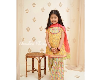 Könnte beinhalten: Ein junges Mädchen trägt ein gelbes und rosafarbenes Salwar Kameez mit floralem Muster und einem rosafarbenen Dupatta. Das Outfit ist mit aufwendigen Stickereien und einem Saum verziert. Das Mädchen steht vor einer weißen Wand mit einem Blumenmuster.