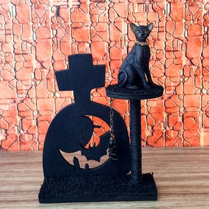 以下が含まれることがあります： 金色の首輪をした黒い猫の置物が小さなプラットフォームに座っています。 コウモリの切り抜きと吊り下げられたチャームが付いた黒い墓石がレンガの壁の前にあります。 ハロウィーンの装飾。