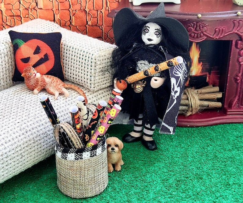 May include: A witch doll wearing a black dress and a black and white cape holds a candy cane wrapped in orange and black paper. The doll is standing on a green surface with a brown and white wicker basket filled with orange, black, and white wrapping paper.