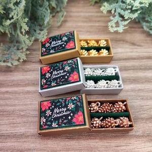以下が含まれることがあります： 黒いラベルに赤いポインセチア、緑の葉、Merry Christmasの文字が描かれた、マッチ箱スタイルの小さな長方形のギフトボックス3つ。各ボックスには、ゴールド、シルバー、コッパーカラーの松ぼっくりの形をした金属製のチャームが入っています。