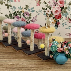 Può includere: Una collezione di tiragraffi per gatti in miniatura con cime colorate e soffici nei toni del rosa, blu e verde. Ogni palo ha una base avvolta in corda e un pompon pendente. Una piccola statuina di gatto è seduta sopra uno dei pali. Un vaso blu con fiori in primo piano.