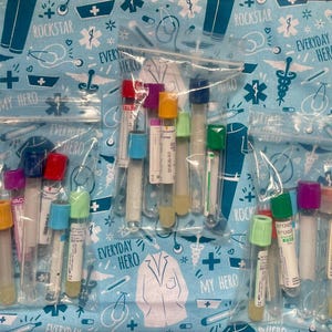 Könnte beinhalten: Mehrere transparente Plastiktüten gefüllt mit medizinischen Reagenzgläsern mit farbigen Kappen. Die Röhrchen enthalten gelbe Flüssigkeit. Der Hintergrund ist ein hellblauer Stoff mit medizinischen Illustrationen und dem Text "Everyday Hero".