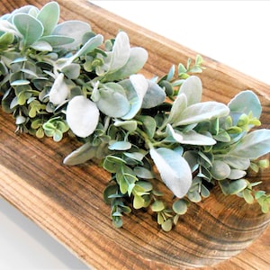 May include: A wooden, oblong-shaped bowl filled with a garland of faux greenery. The garland features soft, silvery-green leaves and smaller, rounder green leaves. The bowl has a natural wood grain finish.