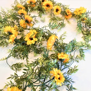 May include: A garland of artificial sunflowers and green leaves. The yellow sunflowers have dark centers and are interspersed with small green leaves. The garland is arranged on a white surface.