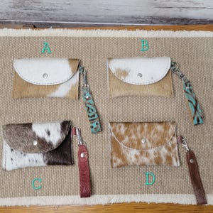 May include: Four cowhide wallets with wrist straps are displayed. Each wallet has a different pattern and color combination. The wallets have a flap closure with a silver snap. The wrist straps are in various colors and designs.
