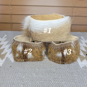 May include: Three cuff bracelets made of textured animal hide with fur. Bracelet #1 features a white and tan gradient pattern. Bracelets #2 and #3 display a brown and white spotted pattern. Each cuff has a smooth tan leather interior lining.