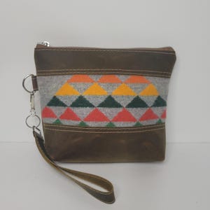 May include: A brown leather clutch with a colorful geometric design. The clutch features a gray wool panel with orange, yellow, green, and red triangles. It has a wrist strap and a silver zipper.