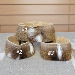 May include: Three circular bracelets made of brown and white animal fur with a smooth tan leather interior lining. Each bracelet features a visible black snap button closure and is labeled with a white number: #1, #2, and #3 on the fur surface.