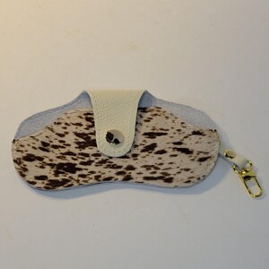 May include: A cowhide eyeglass case with a cream-colored leather flap and a gold-colored clasp. The case features a pattern of brown spots on a cream background. The case is designed to hold eyeglasses.