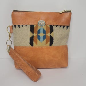 May include: A tan leather and woven fabric clutch with a wrist strap. The clutch features a geometric pattern in black, blue, and tan. The wrist strap is embossed with the word "Rodeo". The clutch has a gold zipper and a silver-toned button.