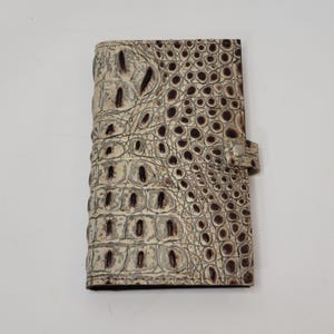 May include: A cream-colored, faux crocodile-skin wallet with a textured surface and a snap closure. The wallet has a rectangular shape and features dark brown accents within the embossed pattern.