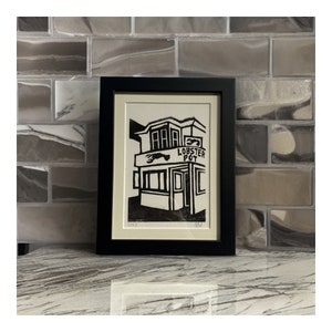 May include: Black and white linocut print of a building with a sign that reads "Lobster Pot" and a lobster illustration. The print is framed in a black frame and is hanging on a wall.
