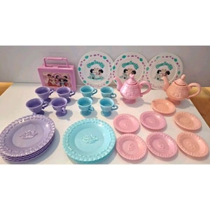 Puede incluir: Un juego de té vintage para niños en colores pastel. El juego incluye platos, tazas, platillos, teteras y una lonchera. Los platos tienen un diseño floral y la lonchera presenta una imagen de dibujos animados. El juego de té es de plástico.