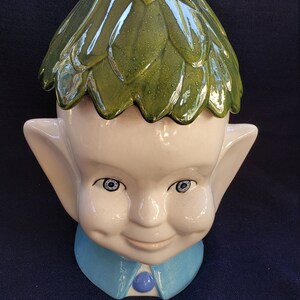 May include: A whimsical ceramic cookie jar shaped like a smiling elf. The jar features a white face with blue eyes, large ears, and a blue collar. The elf's head is covered with a glossy green leaf-like design. A string is tied around the top.