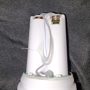 May include: A white cylindrical device with a rounded base, revealing internal components. Two white cylindrical battery compartments are visible, connected by gray wiring. A small spring and electrical components are also present.