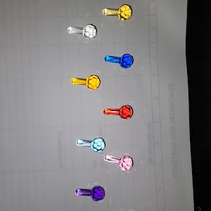 May include: An assortment of colorful, small, decorative glass knobs. The knobs are in various colors, including gold, clear, blue, red, light blue, pink, purple, and green. Each knob has a round, faceted head and a small, elongated stem.