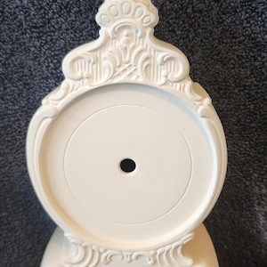 May include: White ceramic clock base with ornate details and a round center opening. The base has a decorative design with a raised center and three legs.