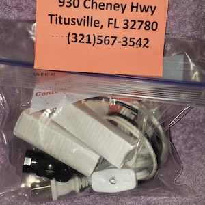 May include: A clear plastic bag containing a light kit with two white light fixtures, a white cord with a black plug, and a white switch. The bag is labeled "LIGHT KIT #2". A pink business card with the text "B 'n' S Ceramics 930 Cheney Hwy Titusville, FL 32780 (321)567-3542" is visible in the background.