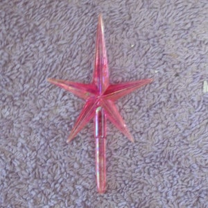 May include: A pink iridescent plastic star tree topper. The star has five points and a long, thin stem.