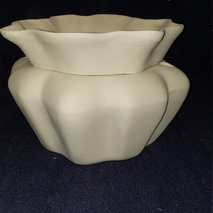 May include: A two-piece, off-white ceramic decorative bowl set. The top bowl has a ruffled edge, and it sits atop a larger, rounded base with a similar fluted design. The set is unpainted and ready for customization.