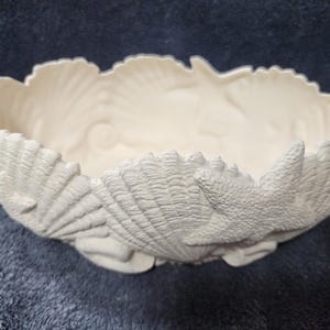May include: Off-white ceramic bowl with a scalloped rim. The bowl is decorated with seashell and starfish designs. The bowl is a decorative piece, suitable for coastal or nautical themed decor.