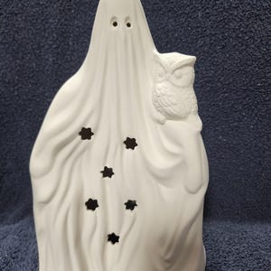 May include: A white ceramic ghost figurine featuring a textured, draped sheet design with two circular eye cutouts. An owl is perched on the ghost's right side, and the front of the ghost is decorated with five small, star-shaped cutouts.