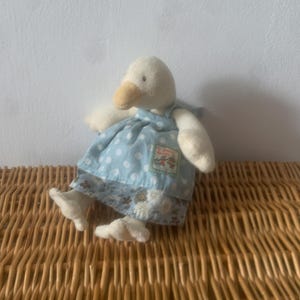 Pu&ograve; includere: Un giocattolo di peluche a forma di oca con corpo color crema, becco giallo e vestito blu a pois. L'abito ha un orlo floreale e una piccola toppa. Il giocattolo &egrave; appoggiato su una superficie di vimini intrecciato.