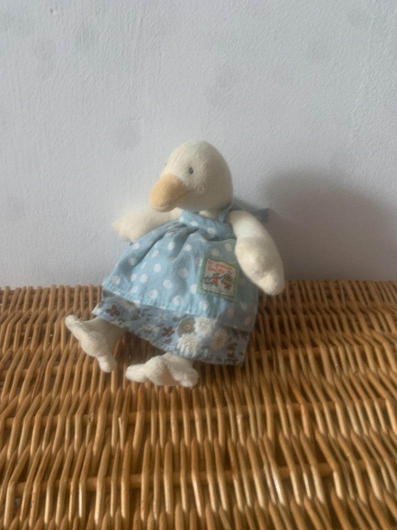 Pu&ograve; includere: Un giocattolo di peluche a forma di oca con corpo color crema, becco giallo e vestito blu a pois. L'abito ha un orlo floreale e una piccola toppa. Il giocattolo &egrave; appoggiato su una superficie di vimini intrecciato.