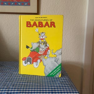May include: A yellow book titled "The Adventures of Babar" by Jean de Brunhoff. The cover features an illustration of Babar and his family in a red car. The book contains three classic stories in one volume.