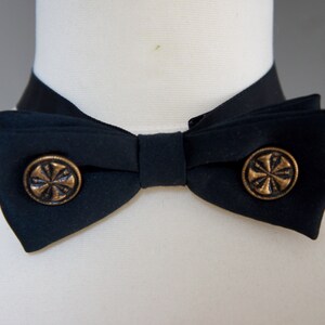 May include: A black bow tie with two gold buttons featuring a stylized cross design.