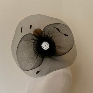 Könnte beinhalten: Ein schwarz-weißer Fascinator-Hut mit einer großen, runden, schwarzen Netzbasis und einer kleineren, runden, weißen Netzmitte. Der Hut ist mit schwarzen Federn und einem weißen Zifferblatt verziert.