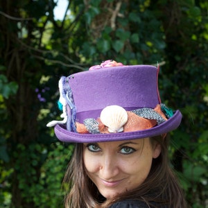 May include: A woman wearing a purple top hat with various embellishments, including seashells and netting. The hat is decorated with a variety of colors, including pink, blue, and orange.