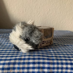May include: A small, gray and white plush dog toy with a black nose and brown eyes. The toy is packaged in a cardboard box with the text "The Dog" on it.