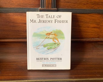 May include: A hardcover book titled "The Tale of Mr. Jeremy Fisher" by Beatrix Potter. The cover features an illustration of a frog fishing on a lily pad. The book is a vintage edition from F. Warne & Co.