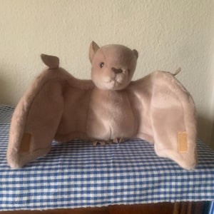 Peut inclure: Une peluche en forme de chauve-souris, de couleur beige et marron, avec les ailes déployées. La chauve-souris a un visage détaillé avec de petites oreilles et un corps doux. Elle est posée sur une surface à carreaux bleus et blancs.