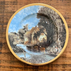 May include: A round decorative plate with a gold rim. The plate features a scenic landscape painting with a house, river, and trees. The sky is blue with white clouds. The plate is set on a wooden surface.
