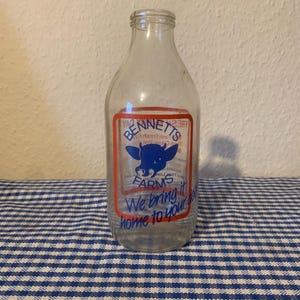 May include: A clear glass milk bottle with a red and blue label. The label reads "Bennetts Farms" with a blue cow graphic and the text "We bring it home to your door."