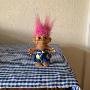 Puede incluir: Un muñeco troll de pelo rosa que lleva un mono azul y sostiene un objeto marrón. El muñeco está de pie sobre una superficie a cuadros azules y blancos.