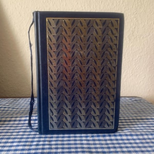 Puede incluir: Un libro azul marino con un patrón geométrico dorado y plateado en la portada. El libro tiene un lomo azul marino a juego y un cordón negro adjunto. El libro está sobre una superficie a cuadros azul y blanco.