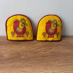 May include: Two yellow oven mitts featuring a red and orange rooster design. The mitts have a maroon trim and are decorated with stylized flowers. The mitts are on a wooden surface.