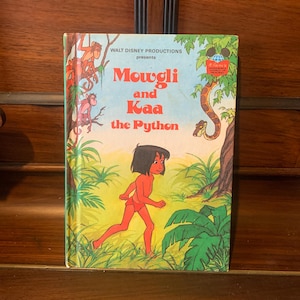 May include: A vintage book cover featuring a jungle scene with Mowgli, a young boy, running through the green foliage. The book title reads "Mowgli and Kaa the Python" and is presented by Walt Disney Productions.