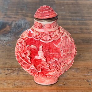 May include: A red and white carved porcelain snuff bottle with a detailed design of figures and flowers. The bottle has a silver lid.