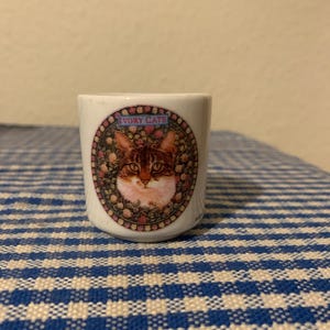 May include: A white ceramic shot glass with a colorful image of a cat's face in a circular frame. The frame has the words "Ivory Cats" in blue lettering.