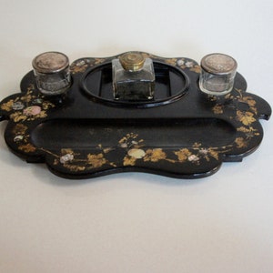 Puede incluir: Un tintero de laca negra con incrustaciones de oro y nácar. El tintero tiene tres compartimentos, uno para tinta y dos para tinteros.