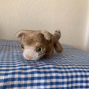 May include: A small, brown and white plush cat toy with green eyes. The cat is lying on a blue and white checkered fabric.
