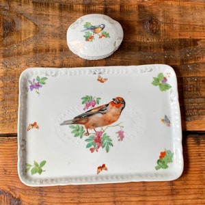Könnte beinhalten: Eine weiße Porzellanschale mit einem zarten Blumendesign und einem Vogel in der Mitte. Die Schale ist mit einem Vogel, Blumen und Schmetterlingen verziert.