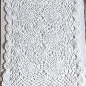 Puede incluir: Doily de ganchillo blanco con un delicado patrón floral. El doily es cuadrado y tiene un círculo central con un círculo más pequeño en el interior.