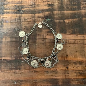 May include: Silver-toned necklace featuring coin pendants and black bead accents. The necklace has multiple layers of chain and is displayed on a wooden surface. The coin pendants have a detailed relief design.